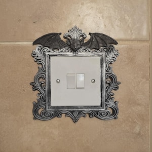 May include: A decorative light switch cover with a bat and ornate frame design. The bat is centered at the top, with wings spread. The frame is silver and surrounds a white light switch with two toggles. The cover is mounted on a beige wall.