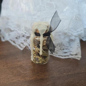 May include: A small glass jar filled with dried herbs and spices, sealed with a wax-covered cork. A black ribbon is tied around the neck of the jar. The jar is set against a backdrop of white lace and a wooden surface.