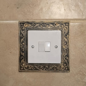 Può includere: Un interruttore doppio bianco con due pulsanti rettangolari, incorniciato in una cornice decorativa. La cornice presenta un design ornato e vorticoso con accenti oro e neri. L'interruttore è montato su una parete piastrellata beige.