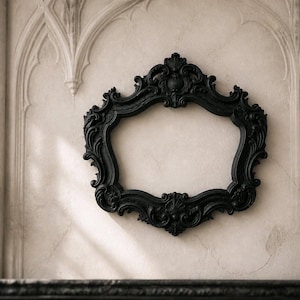 Könnte beinhalten: Ein schwarzer, verzierter Bilderrahmen mit aufwendigen Details hängt an einer strukturierten, cremefarbenen Wand. Links befindet sich ein schwarzer Kerzenhalter mit einer brennenden Kerze. Der Rahmen ist leer.