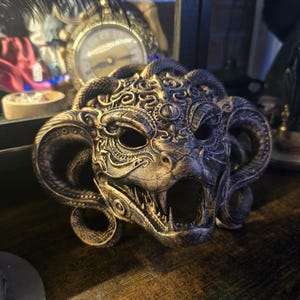 Op de afbeelding: Een gedetailleerd metalen masker met een demonisch ontwerp. Het masker heeft hoorns, slangenachtige details en een open mond met zichtbare tanden. Het kleurenschema is een mix van zilver en brons, met ingewikkelde patronen.