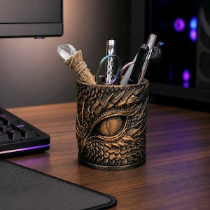 Peut inclure: Un pot à crayons de couleur bronze avec un motif d'œil de dragon. Le porte-crayons cylindrique contient des stylos, des ciseaux et une baguette de cristal. Le support est fait d'un matériau texturé et repose sur un bureau en bois.