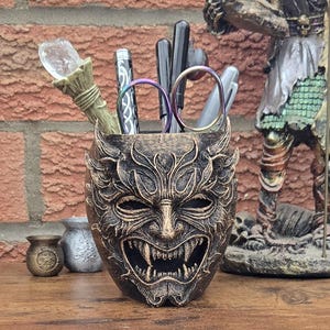 Puede incluir: Un portalápices de color bronce con forma de máscara demoníaca con dientes afilados y detalles intrincados. Contiene bolígrafos, tijeras y una varita decorativa. Una figura y pequeñas macetas se ven en el fondo.