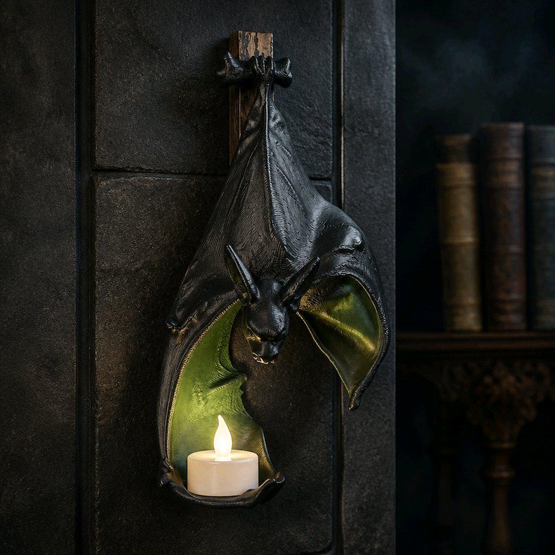 May include: A black bat-shaped wall sconce with a green interior and a small, flickering LED candle. The bat is mounted on a wooden plank and hangs against a dark, textured wall. The wings are spread, and the bat's face is detailed.