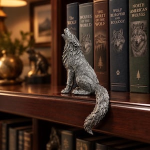 May include: A silver-toned wolf figurine depicted in a howling pose with its head tilted upwards. The sculpture features detailed fur texture and is positioned on the edge of a wooden shelf, with its tail hanging down over the front edge.