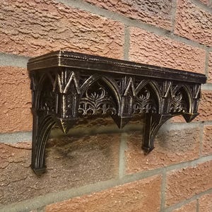 Peut inclure: Étagère murale décorative de style gothique noir et or, fixée sur un mur de briques rouges. L'étagère présente des détails en forme d'arche et des supports pointus. Idéale pour la décoration intérieure.