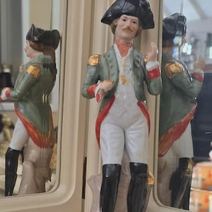 May include: A ceramic figurine depicting a man in a military uniform. The figure wears a black hat, a green jacket with gold details, white trousers, and black boots. The base is white with the letter "N".