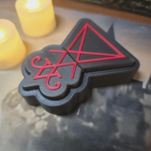 May include: A black and red decorative box with a geometric design. The box has a triangular top with a red pentagram and a stylized symbol. Two yellow candles are in the background.
