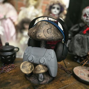 May include: A decorative headphone stand in the shape of a mushroom, holding a grey PlayStation 5 controller and black headphones. The stand is painted in brown and gold, with star and crescent moon details. Two small mushroom accents are at the base.