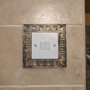 May include: A white double light switch with two rectangular buttons, set within a decorative bronze-colored frame. The frame features a repeating floral pattern. The switch is mounted on a beige tiled wall.