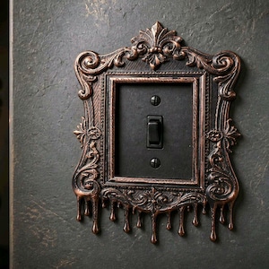 May include: A light switch cover plate featuring an ornate, copper-colored frame with intricate scrollwork and a dripping effect at the bottom. The central rectangular section is matte black with a single black toggle switch and two black screws.