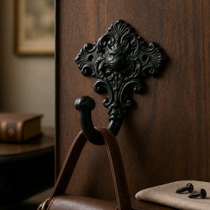 May include: A matte black wall hook featuring an ornate, symmetrical scrollwork design with a central lion head motif. The hook is mounted on a dark wood surface and supports a brown leather strap. Two small metal screws are visible on a nearby cloth.