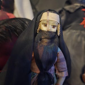 May include: A vintage doll in traditional Middle Eastern dress. The doll wears a black veil, a head covering with gold detailing, and gold earrings. The doll's dress has a colourful striped pattern. A collectible item.