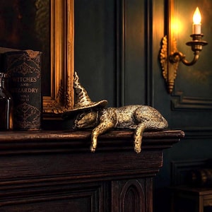 May include: A gold-toned cat figurine wearing a pointed witch hat, resting in a prone position with its front and back legs hanging over the edge of a dark wooden surface. The cat has a textured, metallic finish and a pointed, slightly slumped hat.