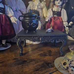 Peut inclure: Une table noire ornée avec des pieds incurvés supporte un chaudron noir avec un couvercle. Un petit objet décoratif est posé sur la table. L'arrière-plan présente plusieurs poupées anciennes en robes.