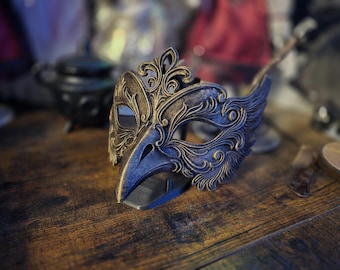 May include: A bird-shaped masquerade mask featuring a pointed beak and intricate, raised scrollwork patterns. The mask is finished in a dark, weathered charcoal grey with metallic gold highlights accentuating the detailed filigree carvings.