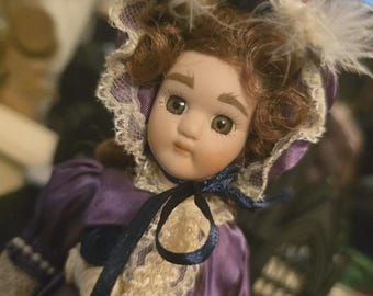 Puede incluir: Muñeca de porcelana con cabello rizado castaño y ojos marrones. La muñeca viste un vestido de satén morado con ribete de encaje y un gorro a juego adornado con plumas. Una cinta azul oscuro está atada alrededor del cuello.