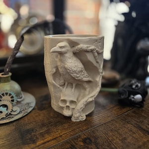 Peut inclure: Un récipient cylindrique beige avec un corbeau sculpté perché sur un crâne. Le corbeau a des plumes détaillées et un bec pointu. Un serpent s'enroule autour du corbeau et du crâne. Le récipient est probablement destiné à contenir des crayons ou de petits objets.