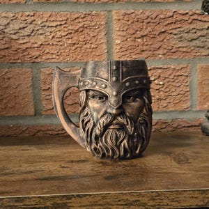 Peut inclure: Une tasse de couleur bronze représentant un guerrier viking avec un casque et une barbe. La poignée est en forme de hache. La tasse est conçue pour les boissons et est fabriquée en matériau métallique.
