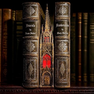 May include: Two antique-style books titled Dracula by Bram Stoker and Frankenstein by Mary Shelley, featuring dark green leather-like covers with silver embossed details, flanking a central miniature gothic tower structure with glowing red windows.