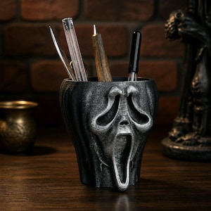 Op de afbeelding: Een zwarte en zilveren pennenhouder in de vorm van het Scream-masker, gevuld met pennen en potloden. De pennenhouder staat op een houten oppervlak, met een bakstenen muur op de achtergrond. De pennenhouder heeft een textuur.