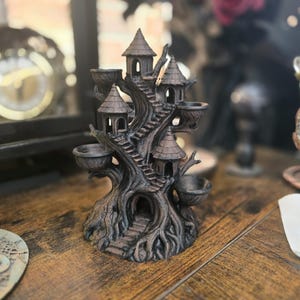 May include: A detailed, dark brown tree-shaped incense holder with multiple small tower-like structures and bowls. The design includes stairs and a hollowed-out base, creating a whimsical, gothic aesthetic. The holder sits on a wooden surface.
