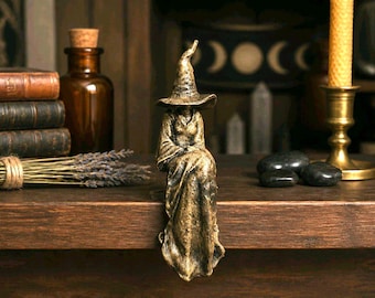 May include: A bronze-toned figurine of a seated witch wearing a tall, pointed hat and a long, flowing robe. The figure is crafted with a textured, weathered finish and is positioned to sit on the edge of a flat, horizontal surface.