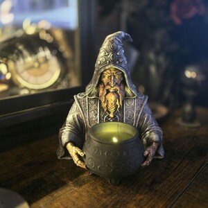 May include: A detailed figurine of a wizard holding a cauldron with a lit candle. The wizard is dressed in a silver robe and pointed hat, with a long, golden beard. The cauldron is black with ornate detailing. The background is blurred, with hints of a clock and other decor.