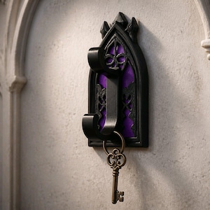 May include: A black wall hook featuring an arched frame with intricate cutouts and a vibrant purple backing. The hook has two curved prongs, with a metallic antique brass-colored skeleton key hanging from the lower prong against the wall.