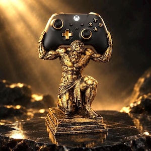 May include: A gold-toned statue of a kneeling figure with a muscular physique, supporting a black and gold wireless game controller above its head. The figure is positioned on a rectangular base, featuring detailed textures and a weathered finish.