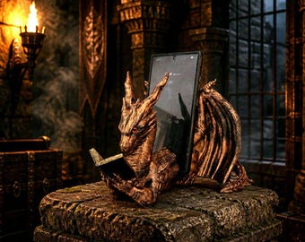 May include: A bronze-colored dragon figurine with red eyes, holding a small open book in its claws. The dragon features detailed scales and folded wings, and it serves as a stand for a rectangular smartphone placed upright behind its back.