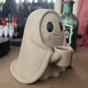 May include: A small, light beige ghost figurine draped in a textured blanket with a repeating circular pattern. The ghost has large, round black eyes, a simple curved smile, and is holding a small, light beige mug filled with a brown liquid.