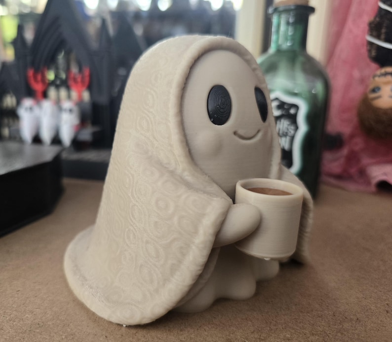 May include: A small, light beige ghost figurine draped in a textured blanket with a repeating circular pattern. The ghost has large, round black eyes, a simple curved smile, and is holding a small, light beige mug filled with a brown liquid.