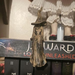 Puede incluir: Una figura de color bronce de un personaje sentado que lleva una t&uacute;nica larga y fluida y un sombrero puntiagudo. La figura est&aacute; colocada en el borde del lomo de un libro negro, con el texto J. WARD y UNLEASHED visible en la cubierta.