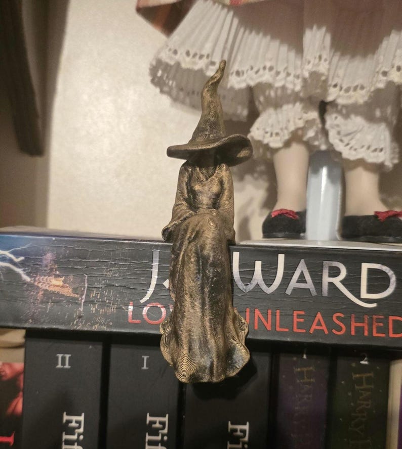 Puede incluir: Una figura de color bronce de un personaje sentado que lleva una t&uacute;nica larga y fluida y un sombrero puntiagudo. La figura est&aacute; colocada en el borde del lomo de un libro negro, con el texto J. WARD y UNLEASHED visible en la cubierta.