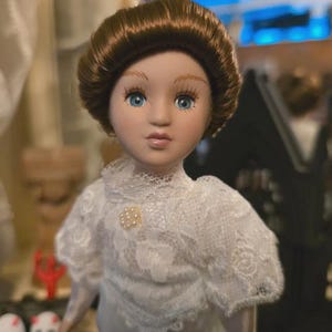 May include: A porcelain doll with blue eyes and brown hair in an updo. The doll wears a white lace dress with a pearl brooch. A collectible doll.