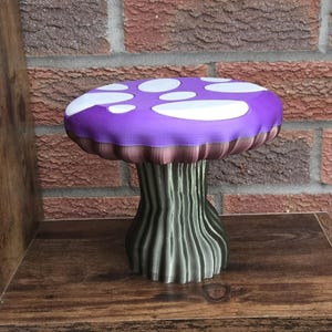 Può includere: Sgabello decorativo a forma di fungo, con una parte superiore viola con macchie bianche. La parte inferiore del cappello è marrone e il gambo è verde. Realizzato in materiale stampato in 3D, poggia su una superficie di legno. Altezza: circa 25 cm.