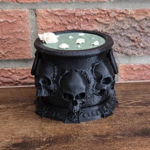 Peut inclure: Un récipient noir en forme de chaudron avec trois motifs de crânes sur le devant. Le chaudron a un intérieur vert avec des décorations blanches en forme de crâne. Le chaudron repose sur une base décorative et possède deux poignées.
