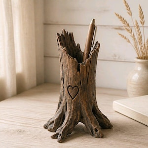 May include: A brown, hollowed-out tree stump pen holder with a textured, bark-like exterior and a carved heart symbol on the front. The stump features a jagged, uneven top edge and a wide, root-like base, containing a single metallic pen.