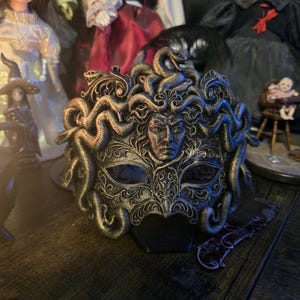 Può includere: Una maschera decorativa di Medusa, nera e dorata, con un volto dettagliato e serpenti. La maschera presenta disegni intricati ed è un oggetto decorativo unico. È un'opera d'arte unica.