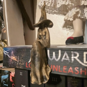 Puede incluir: Una figura de color bronce de una bruja con un sombrero puntiagudo y una t&uacute;nica larga y fluida, representada en posici&oacute;n sentada con las piernas colgando sobre el borde de un libro titulado J.R. Ward Lover Unleashed, con acabado met&aacute;lico.