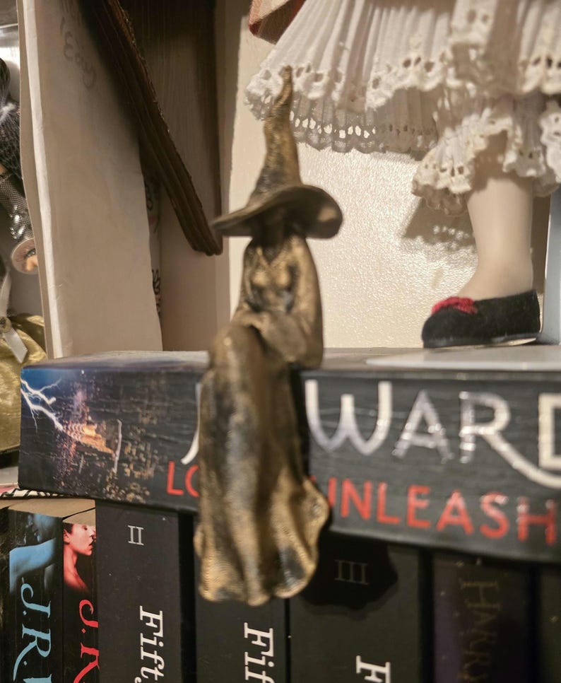Puede incluir: Una figura de color bronce de una bruja con un sombrero puntiagudo y una t&uacute;nica larga y fluida, representada en posici&oacute;n sentada con las piernas colgando sobre el borde de un libro titulado J.R. Ward Lover Unleashed, con acabado met&aacute;lico.