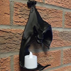 May include: A black wall-mounted candle holder shaped like a bat, with a small white LED candle inside. The holder is mounted on a red brick wall, creating a gothic or Halloween theme.