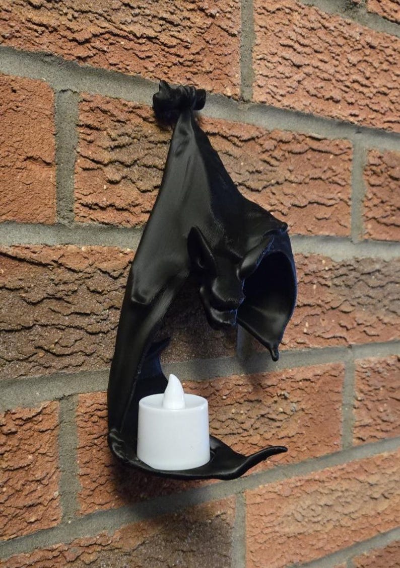 May include: A black wall-mounted candle holder shaped like a bat, with a small white LED candle inside. The holder is mounted on a red brick wall, creating a gothic or Halloween theme.