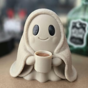 May include: A small, light beige ghost figurine with a smooth, rounded shape, featuring two large, dark, oval-shaped eyes and a subtle, curved smile. The ghost is holding a small, matching beige mug filled with a brown liquid in its hands.