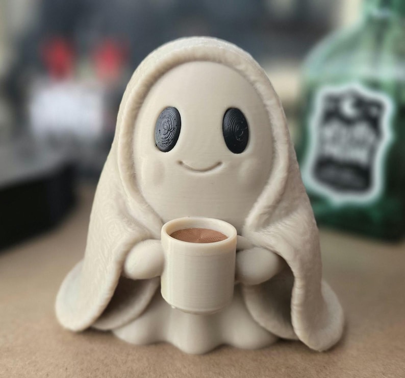 May include: A small, light beige ghost figurine with a smooth, rounded shape, featuring two large, dark, oval-shaped eyes and a subtle, curved smile. The ghost is holding a small, matching beige mug filled with a brown liquid in its hands.