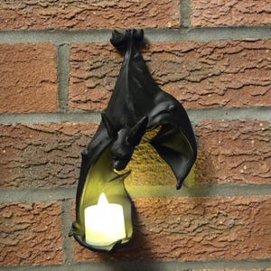 May include: Black bat-shaped wall sconce with a small, white candle inside. The bat is hanging upside down on a red brick wall. The candle provides a warm, yellow glow. A decorative item for Halloween or gothic decor.