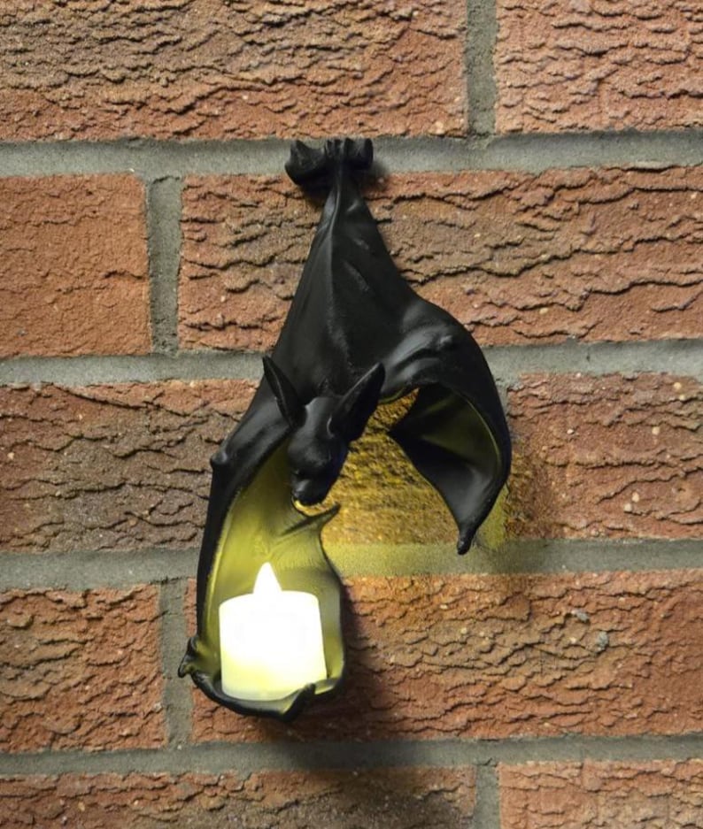 May include: Black bat-shaped wall sconce with a small, white candle inside. The bat is hanging upside down on a red brick wall. The candle provides a warm, yellow glow. A decorative item for Halloween or gothic decor.