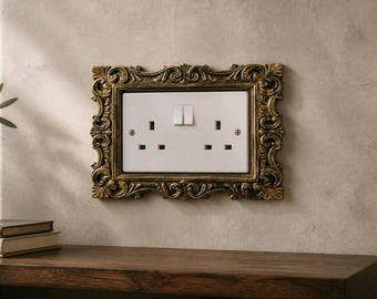 Puede incluir: Un enchufe doble blanco y un interruptor de luz están dentro de un marco dorado ornamentado. El marco está montado en una pared de tono neutro sobre una superficie de madera oscura. El enchufe tiene dos enchufes y dos interruptores.