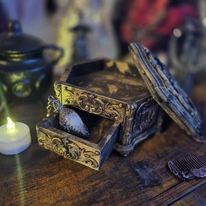 Pode incluir: Uma caixa de madeira em estilo antigo aberta, com entalhes ornamentados, um pingente dentro e um pente decorativo. A caixa é marrom escuro com detalhes dourados. Uma pequena vela acesa está por perto.